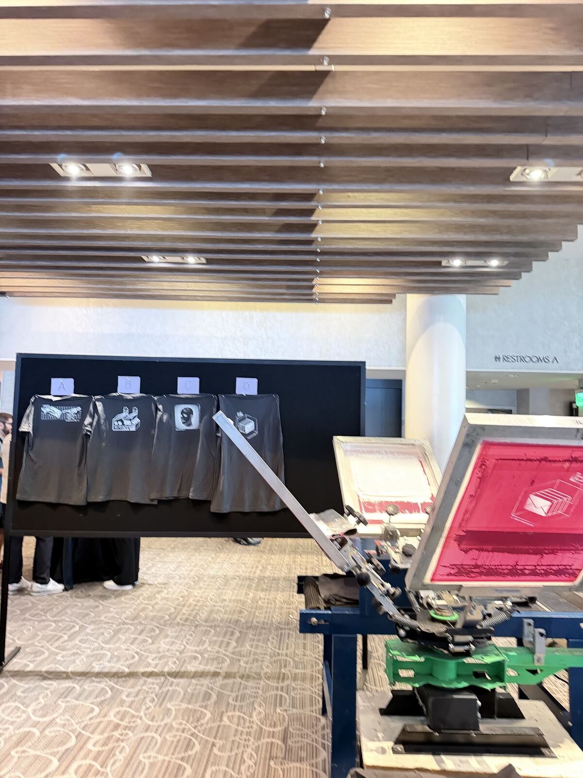 Sample wall of finished designs labeled A through D beside a live press station at a hotel venue