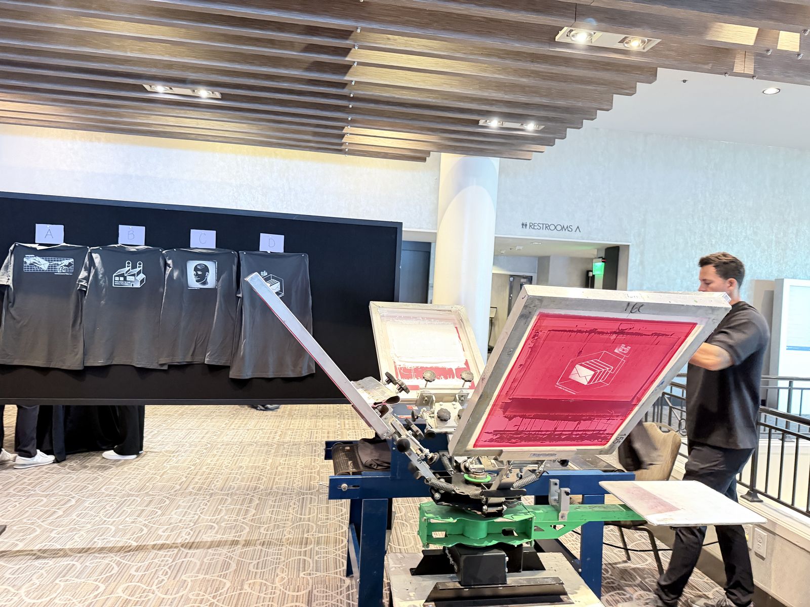 Crew member operating a live print station beside a display wall of finished shirt designs at a hotel conference