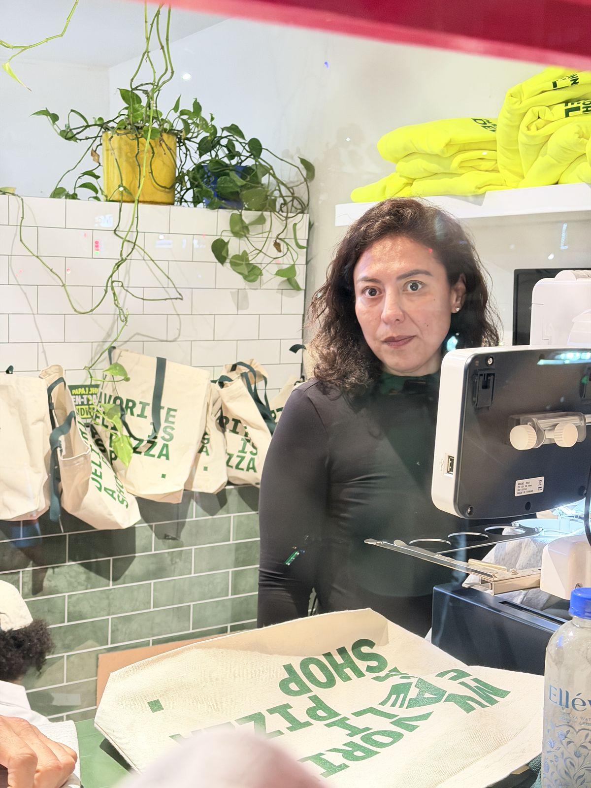 Staff member at a pop-up merch counter with folded neon merchandise stacked on shelves behind her